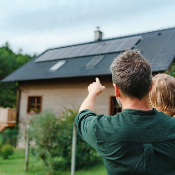 Rear view of dad holding her little girl in arms and showing at their house with solar panels.Alternative energy, saving resources and sustainable lifestyle concept.